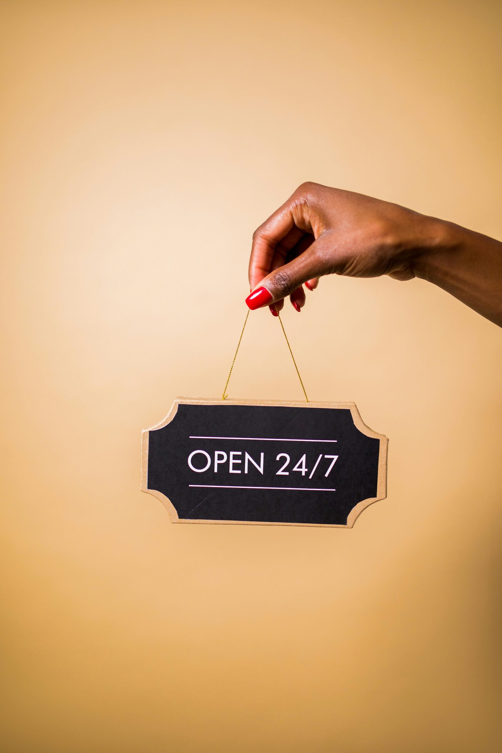A hand with red manicured nails holding a chalkboard sign that reads 'OPEN 24/7' on a neutral background.
